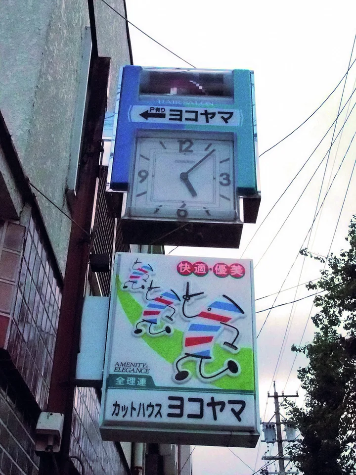 A square clock sits above a colorful barber sign projecting beside a tiled wall and overhead wires.