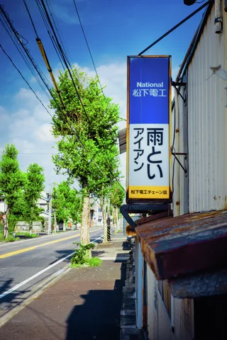 A tall blue and yellow sign projects beside a tree-lined road beneath utility wires and blue sky.