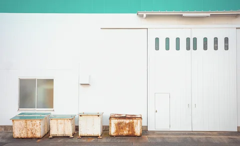 Four rust-stained containers stand against a white industrial wall beside a large door with narrow windows.