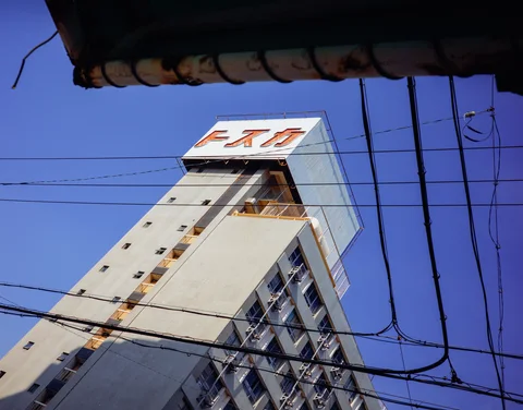 A tall pale building angles upward behind crisscrossing wires beneath blue sky and a dark foreground eave.