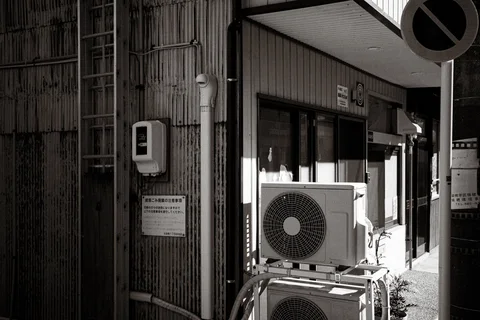 Two air conditioners stand outside a corrugated building beside utility pipes and a street sign in black and white.