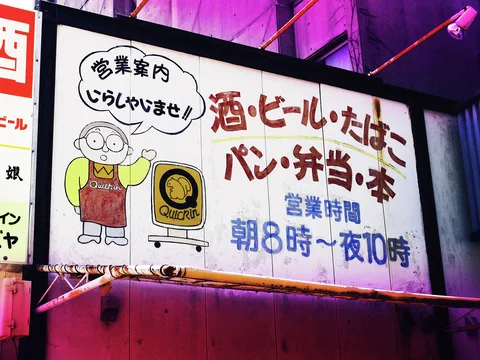 A cartoon shopkeeper and grocery emblem decorate a white sign filled with red and blue Japanese lettering.