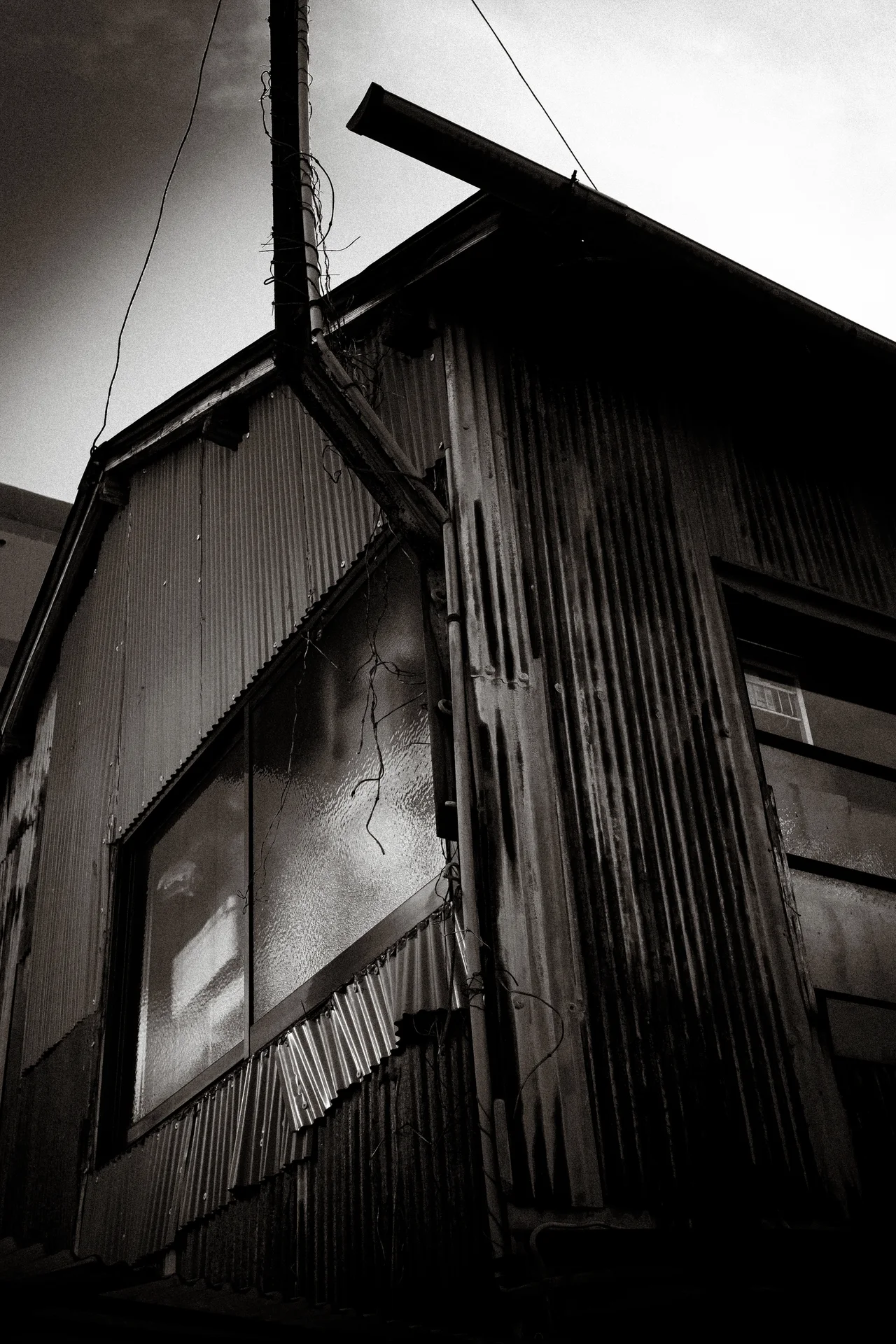 A corrugated building rises beneath crossed roof beams and a utility pole in black and white.