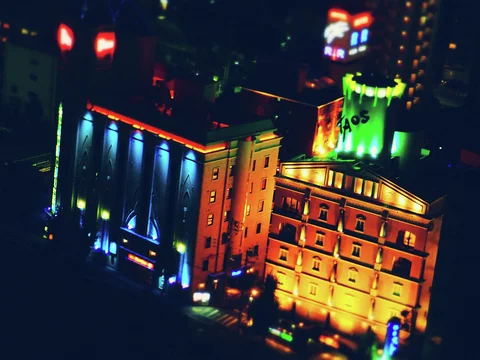 Two buildings glow with vivid blue and orange exterior lighting amid softly blurred city lights.