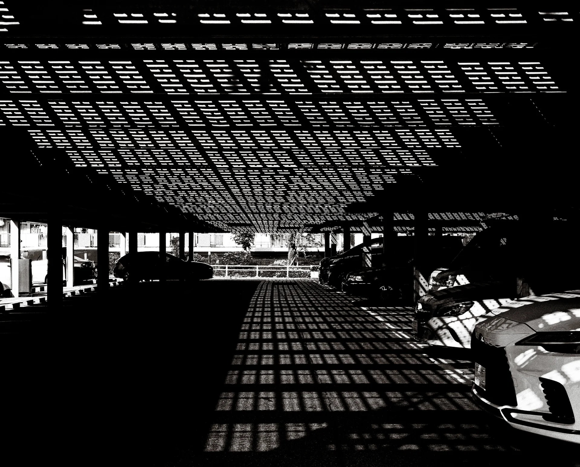 A perforated canopy casts dense rectangular light patterns over parked cars and pavement in black and white.