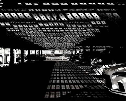 A perforated canopy casts dense rectangular light patterns over parked cars and pavement in black and white.