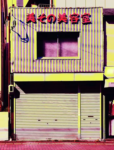 Red Japanese lettering crowns a yellow vertically striped storefront with a dark upstairs window and closed pale shutters.