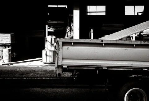 A small flatbed truck catches bright sunlight inside a dark industrial space in black and white.