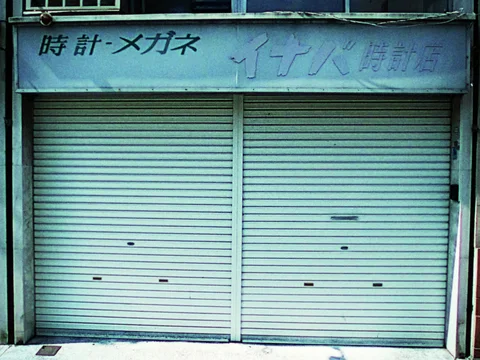 Two closed pale green shutters sit beneath faded Japanese signage on a weathered blue-green storefront.