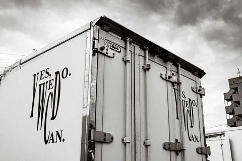 Stylized dark lettering decorates a white box truck beneath textured gray clouds in black and white.