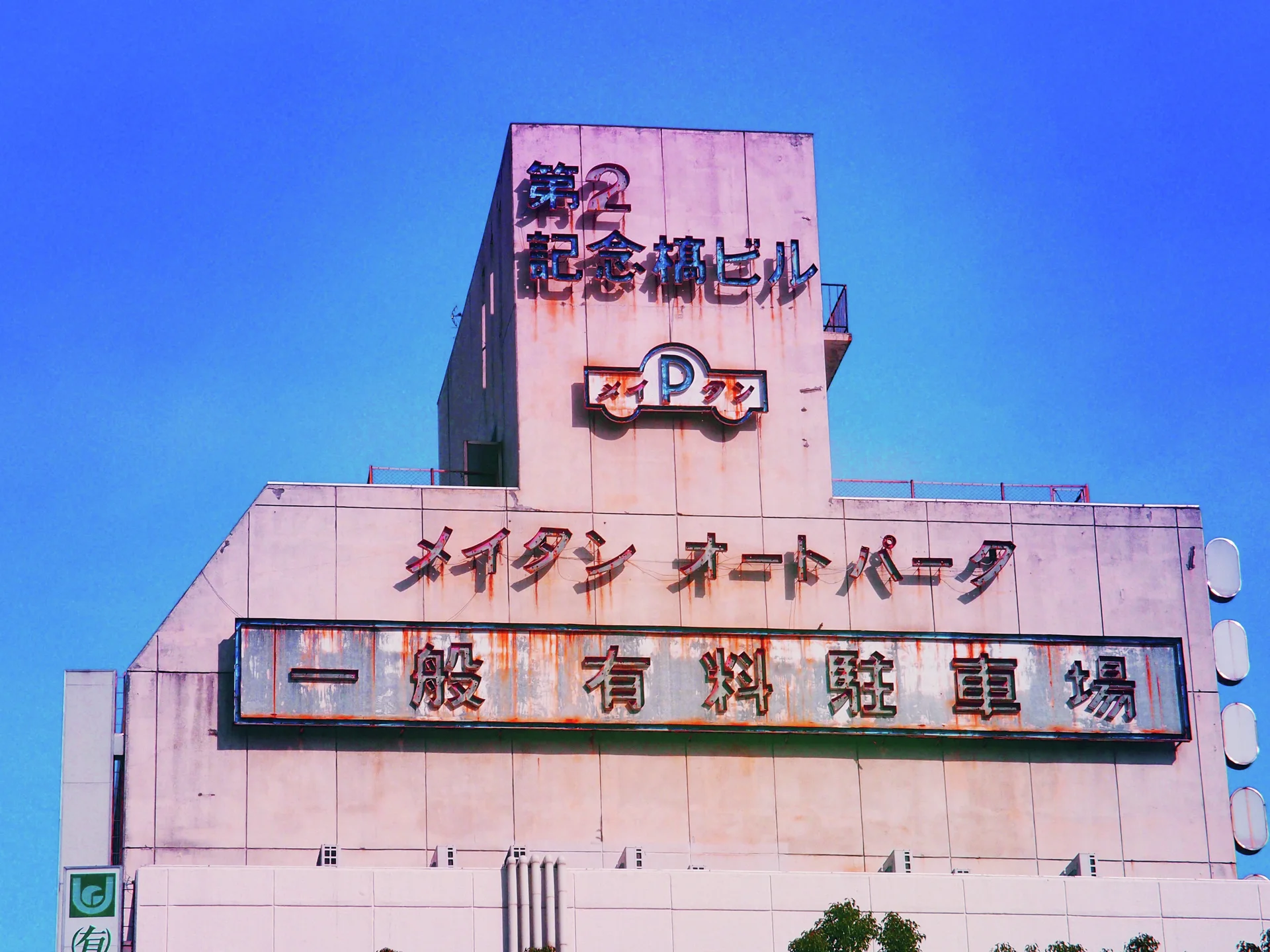Large Japanese signs and a parking emblem cover a pale pink stepped facade beneath vivid blue sky.