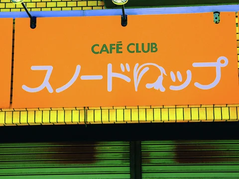 An orange Cafe Club sign with white Japanese lettering hangs above closed green shutters framed by yellow tiles.