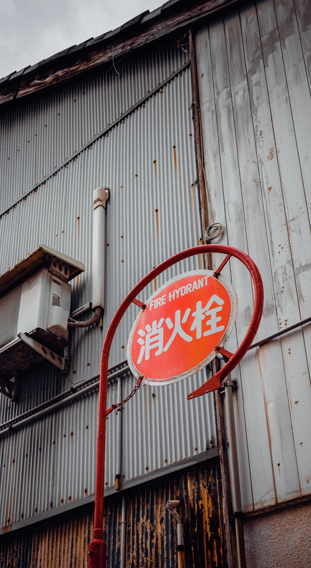 A red oval fire-related sign hangs before gray corrugated siding beside an old wall lantern.