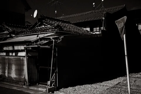 Tiled-roof wooden buildings sit in strong shadow beside a gravel area in black and white.