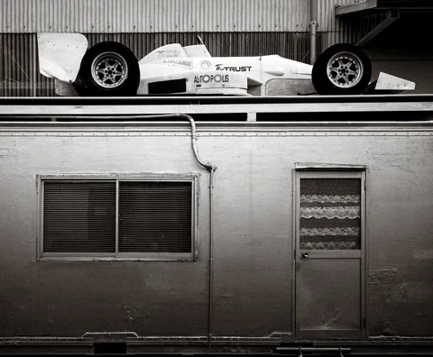 A white open-wheel racing car rests above a plain wall with windows and a door in black and white.