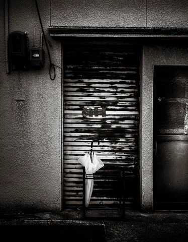 A folded umbrella hangs against a peeling metal shutter beside a dark doorway in black and white.