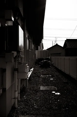 A narrow debris-strewn passage runs beside a dark building toward low rooftops in black and white.