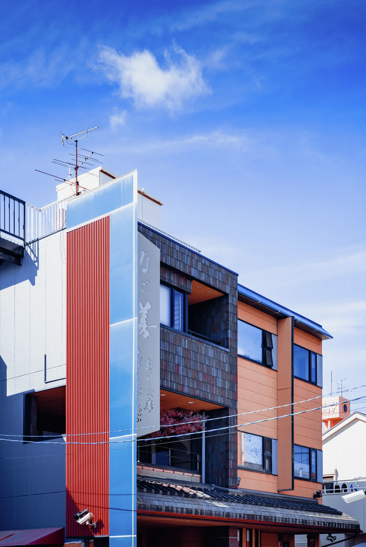 Orange and blue vertical panels frame dark windows on a building beneath a bright blue sky.