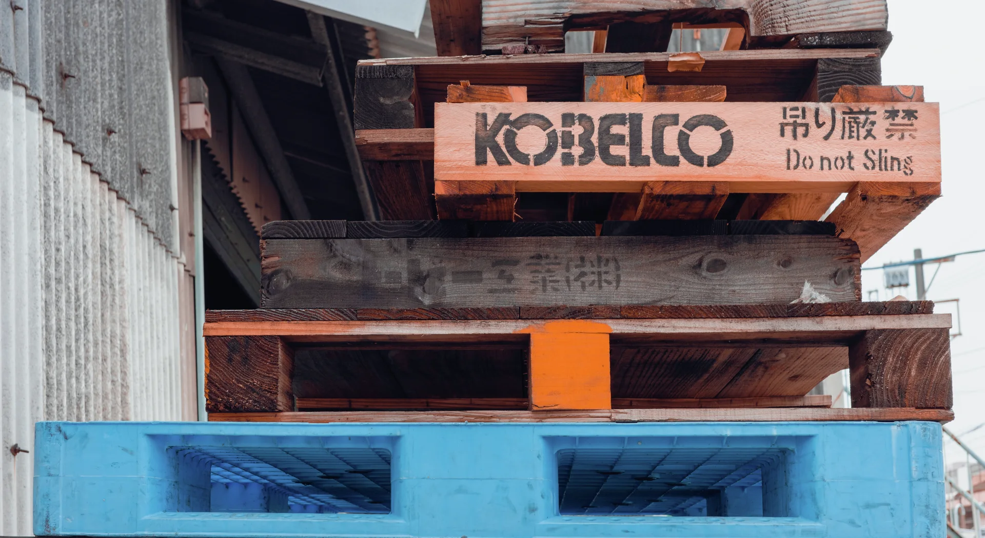Weathered wooden pallets stack above a bright blue plastic pallet beside corrugated warehouse siding.