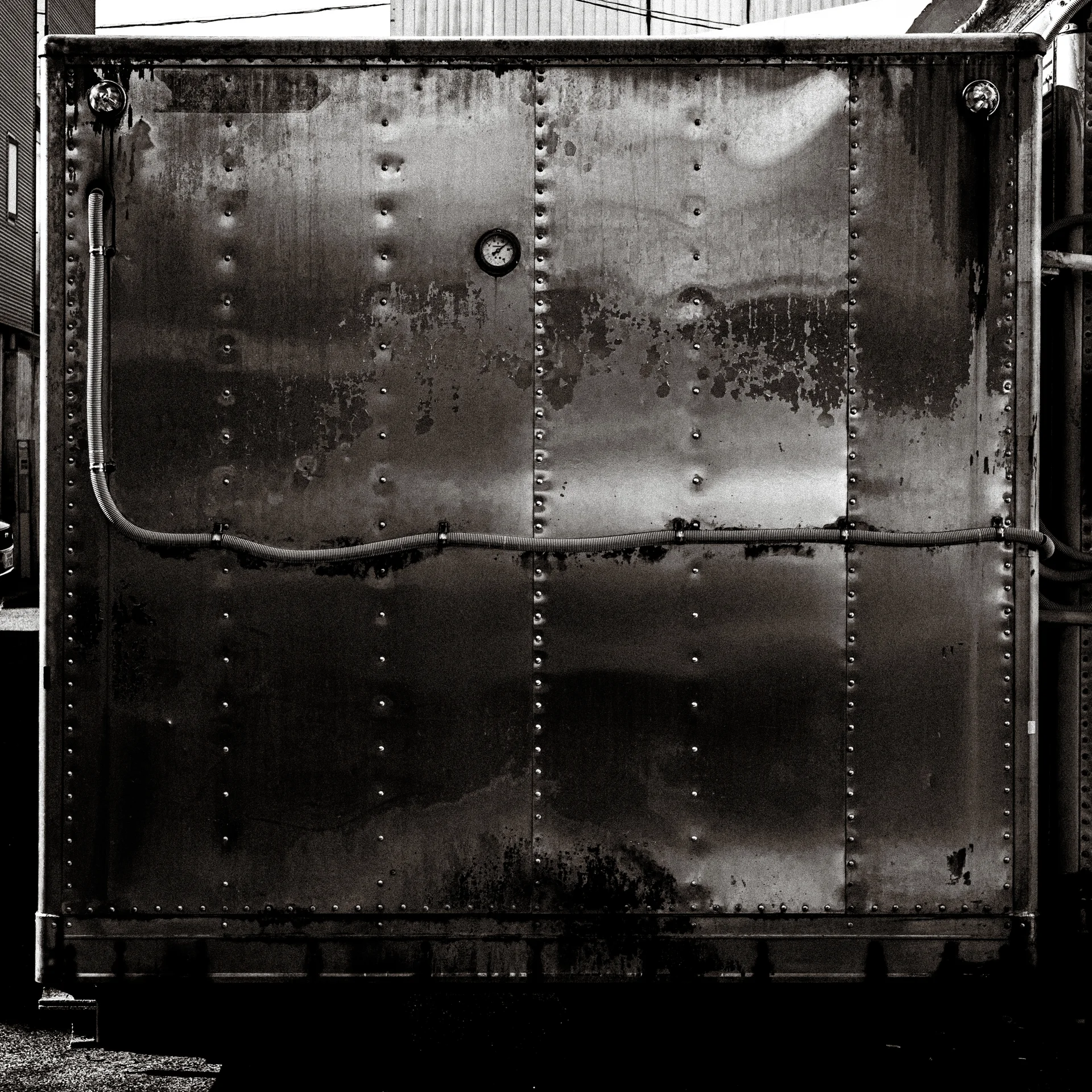 A riveted metal wall with a horizontal cable reflects uneven light in black and white.