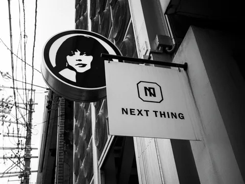 A circular face emblem and pale Next Thing sign project from a building beside overhead wires.