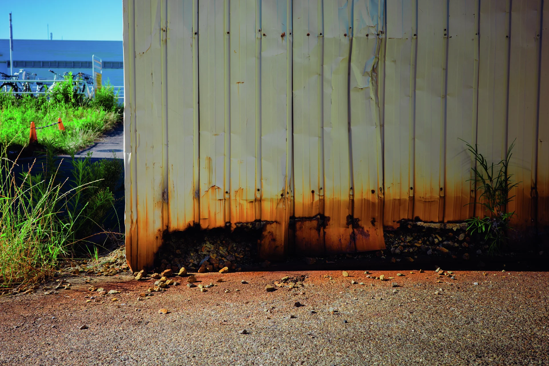 Orange rust stains spread along the base of pale corrugated siding beside weeds and rough pavement.