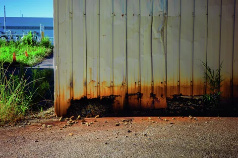 Orange rust stains spread along the base of pale corrugated siding beside weeds and rough pavement.