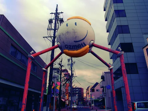 A weathered smiling globe tops red gateway supports above a narrow street between tall buildings.