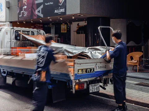 Two people stand beside a small flatbed truck carrying covered materials outside a dark storefront.