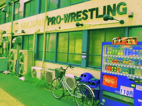 Large green PRO-WRESTLING lettering spans a building above air-conditioning units, a bicycle and a blue vending machine.