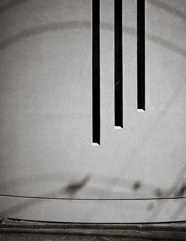 Three narrow vertical recesses punctuate a pale concrete wall above soft shadows and two horizontal wires.