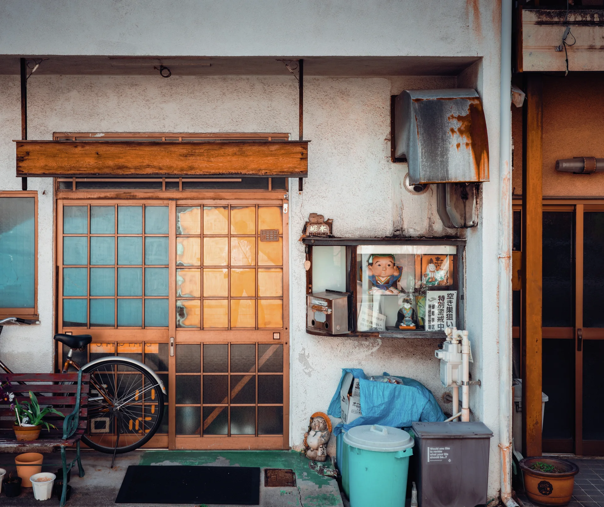A bicycle and plastic bins flank a glazed wooden doorway beneath rusted exterior fixtures.