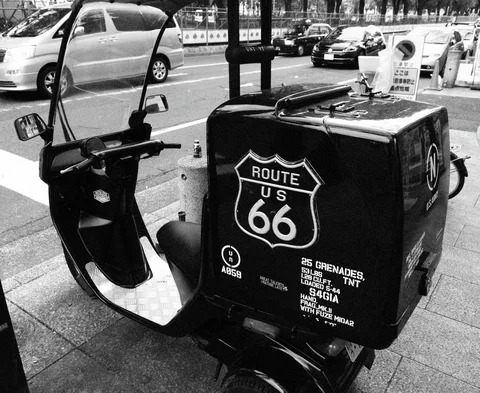 A parked scooter carries a large black cargo box decorated with a Route 66 emblem in black and white.