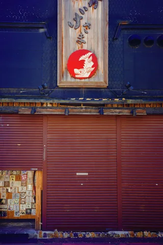 A vertical sign with a red emblem hangs above closed reddish shutters on a blue facade.