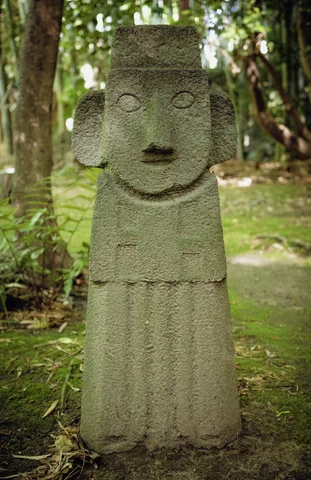 A tall weathered stone figure with a broad nose and carved robe stands among trees.