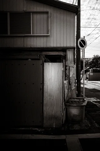 A narrow weathered corrugated house stands beside a roadside sign in black and white.