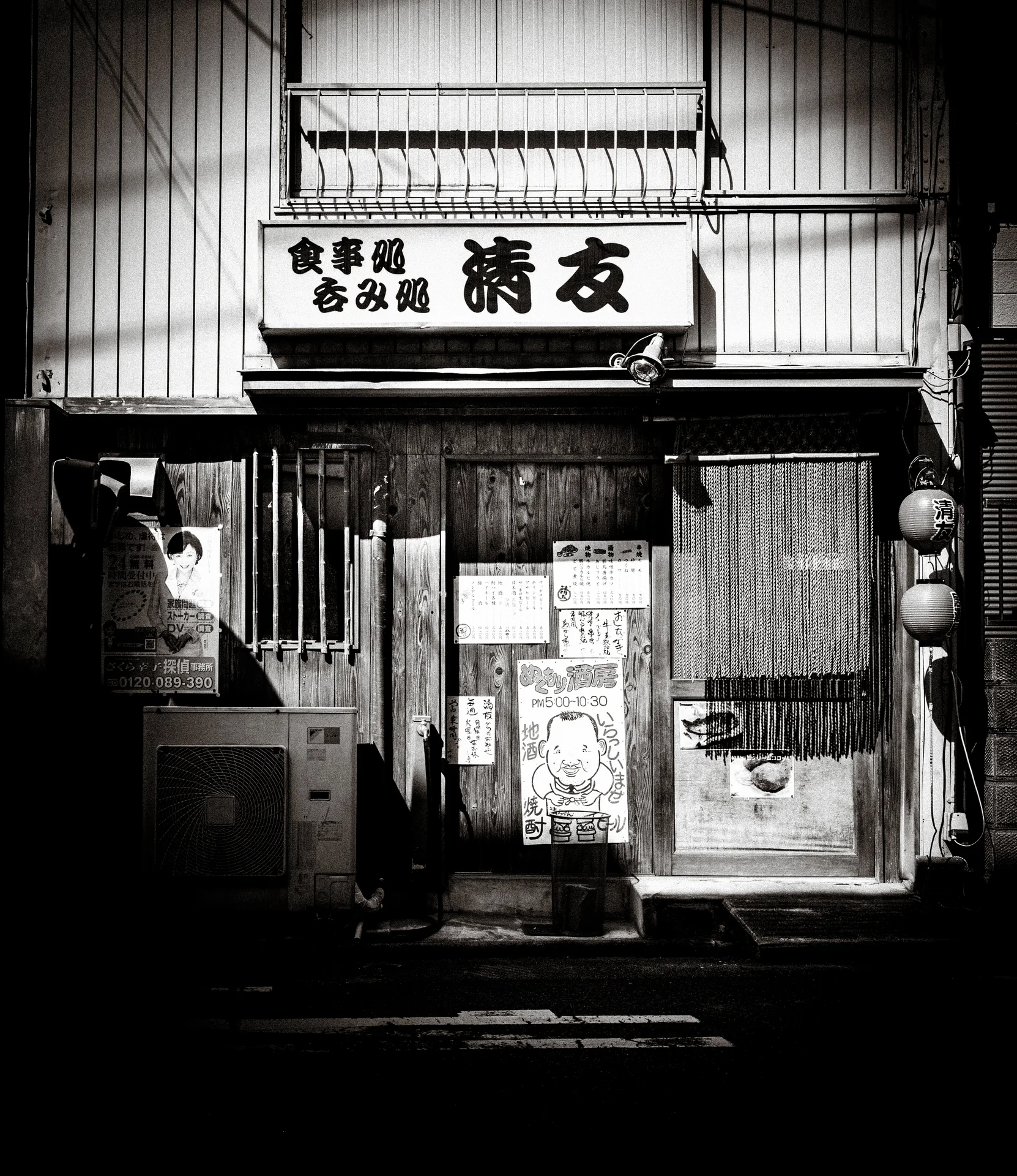 Japanese signage hangs above a weathered storefront entrance covered with notices in a black and white view.