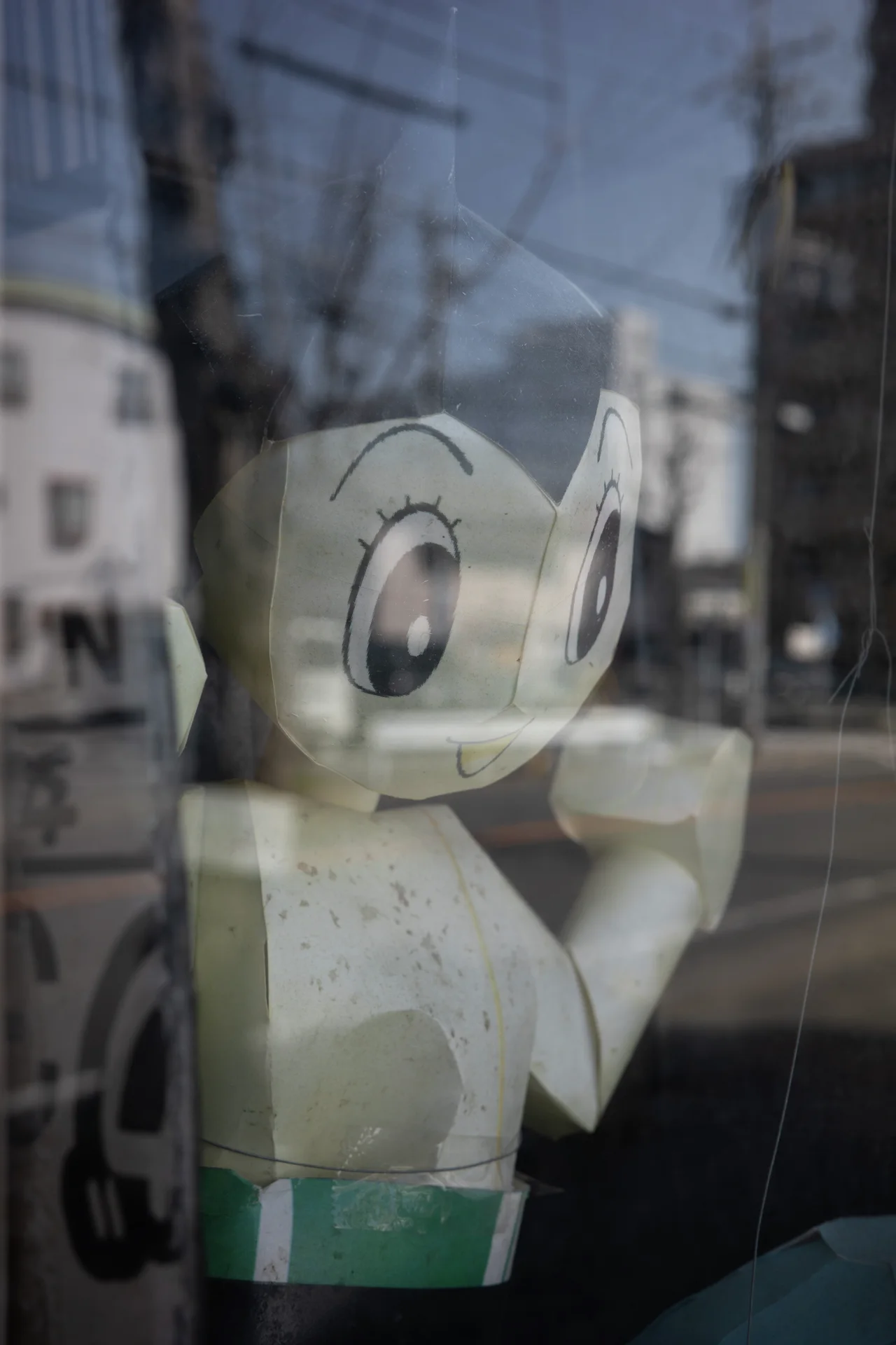 A pale cartoon figure behind glass overlaps street reflections and faint signage in the window.