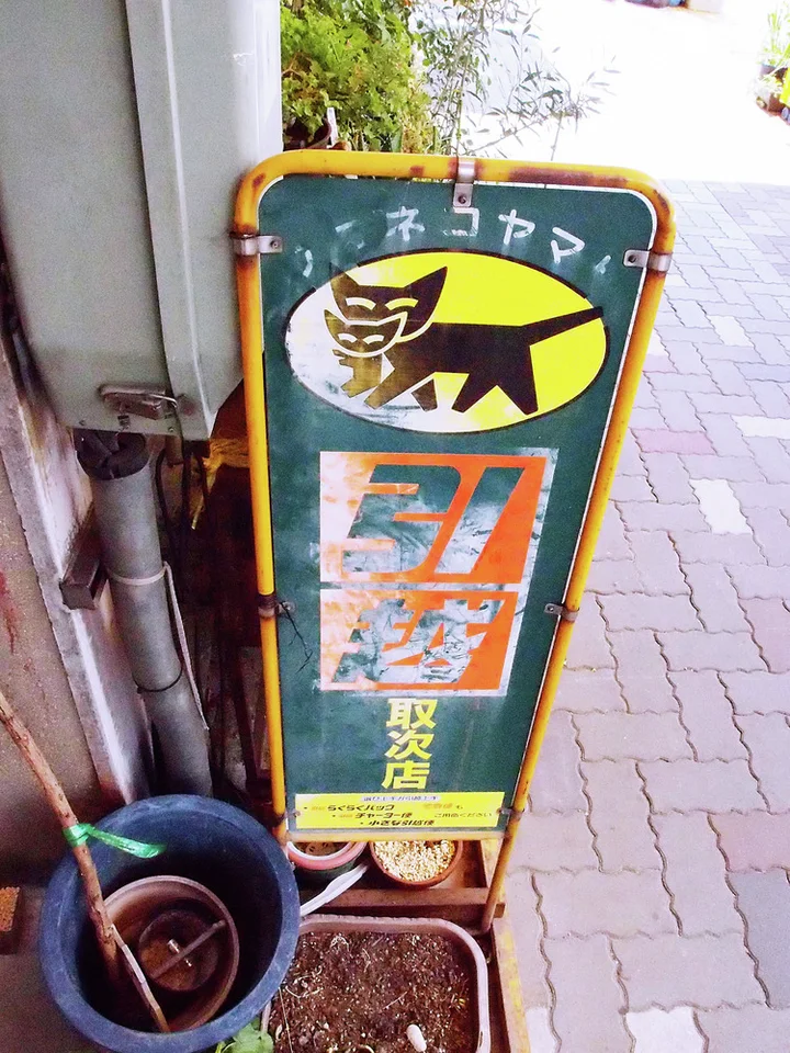A narrow green sign with a cartoon cat leans beside plant pots on a brick-paved sidewalk.