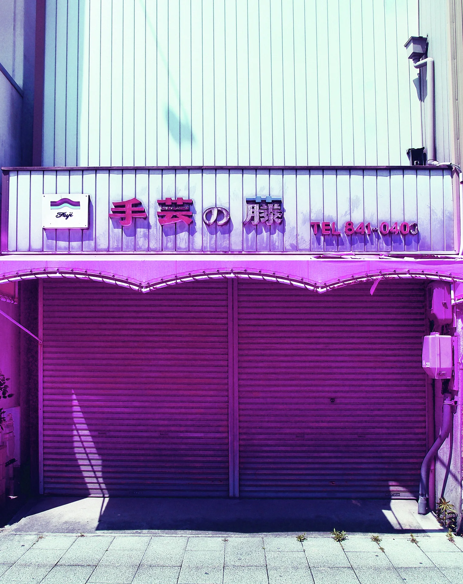 Closed purple shop shutters sit beneath a pale corrugated facade and narrow raised-letter sign.