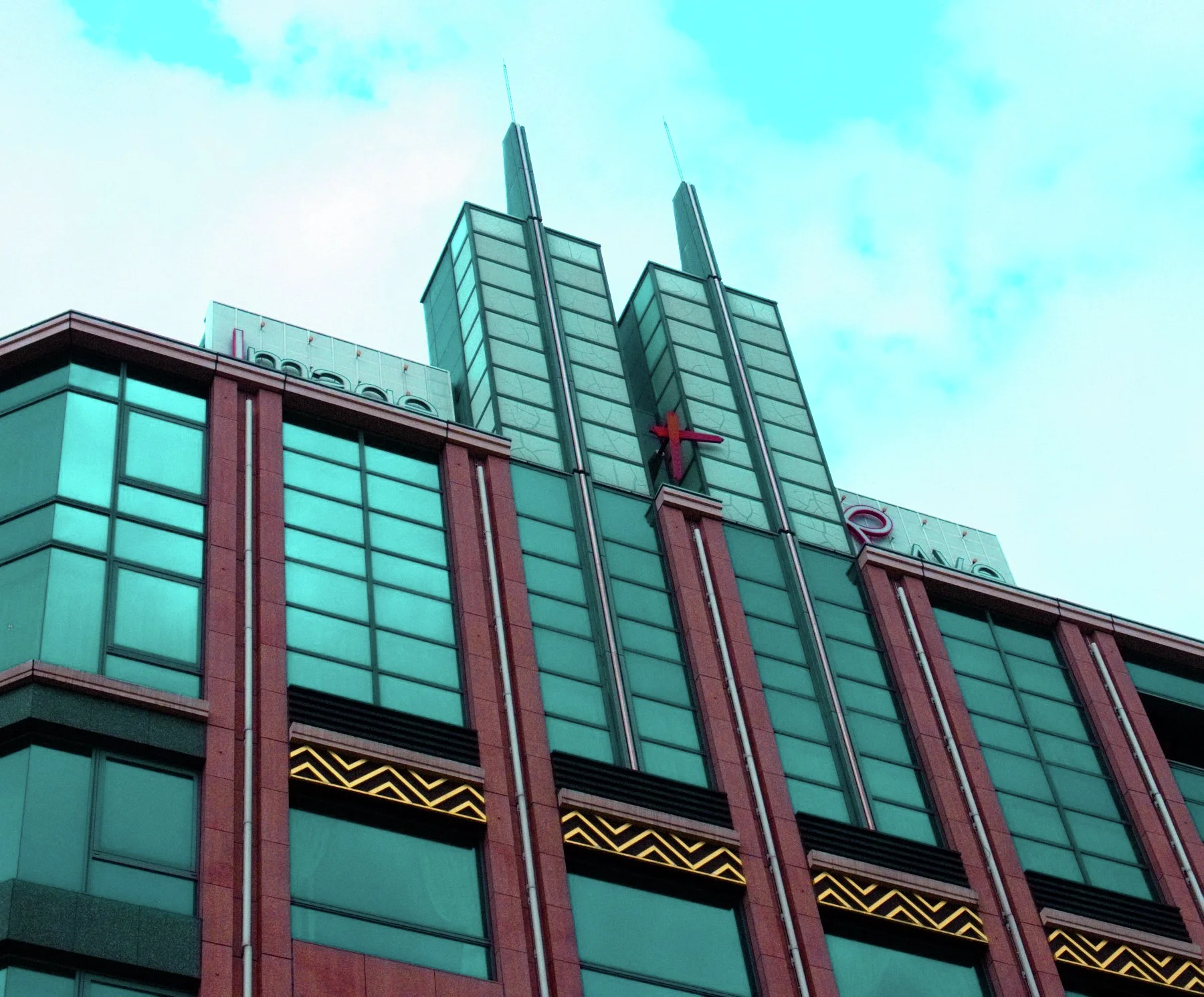 Two pointed vertical towers crown a turquoise-windowed building divided by reddish columns and gold geometric trim.