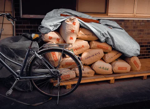 A bicycle stands before stacked beige sacks partly covered by a gray tarp.