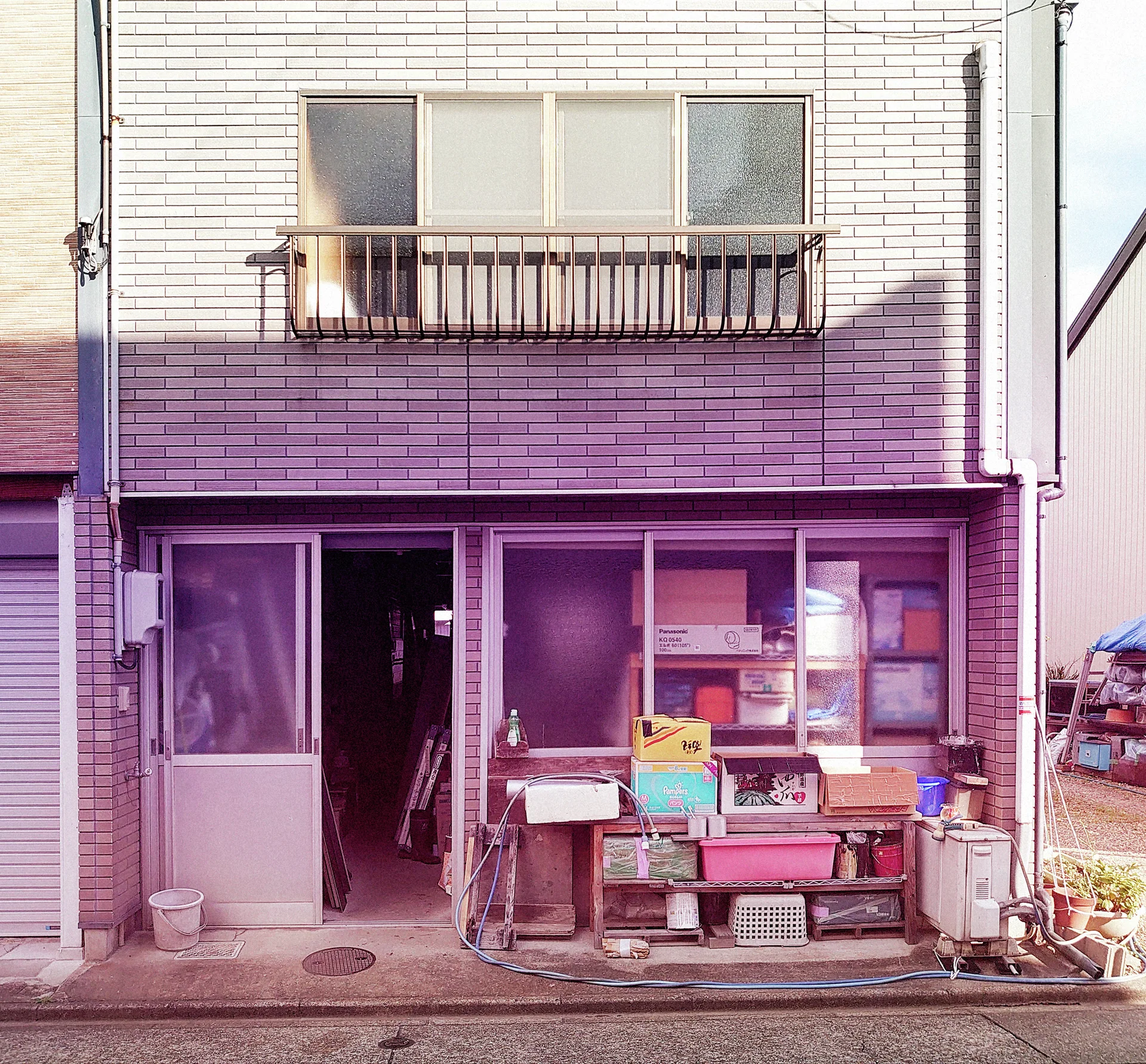 Boxes and household objects crowd a small storefront beneath a pale tiled facade and shallow balcony.