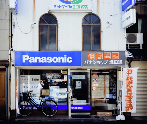 Blue Panasonic signage spans a narrow shop beneath paired arched windows, with a bicycle outside.