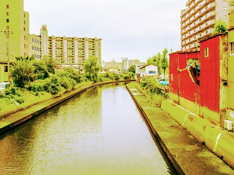 A straight canal runs between green banks and apartment buildings beside a long red industrial wall.