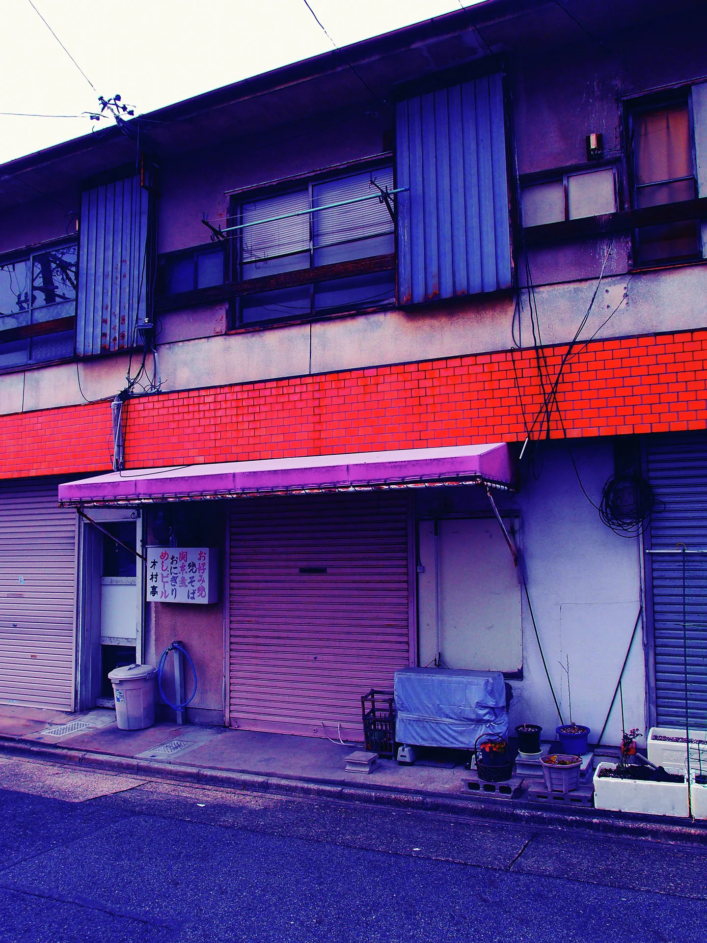 Closed shutters line a low storefront beneath red horizontal tiles and blue upper window panels.