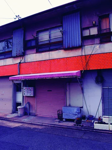 Closed shutters line a low storefront beneath red horizontal tiles and blue upper window panels.