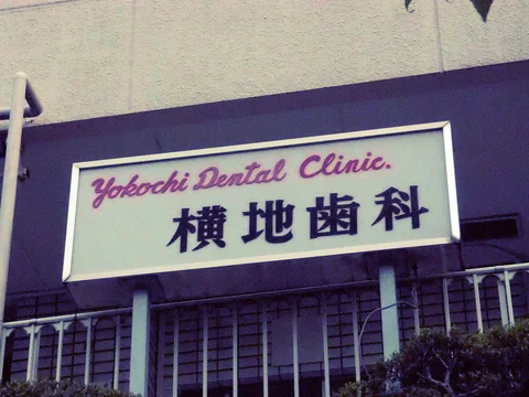 A pale dental clinic sign with pink English script and dark Japanese characters sits above barred windows.