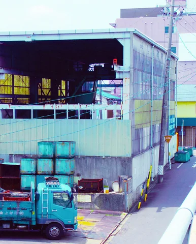 A turquoise truck stands below an open-sided industrial building beside stacks of blue metal containers.