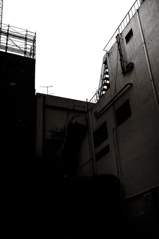 Dark building walls border a narrow alley beneath a bright sky in a black and white view.